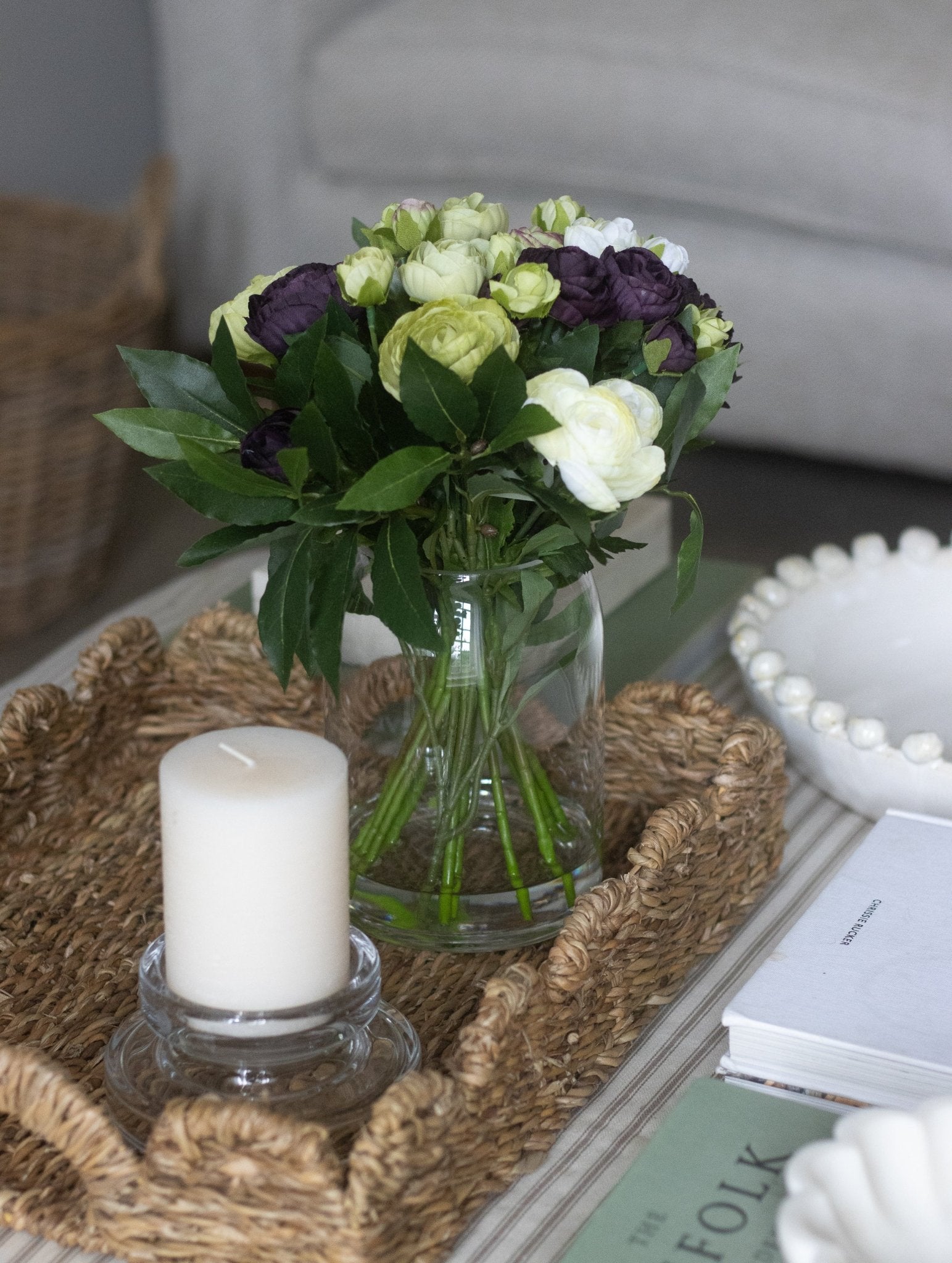 Coffee table close-up with Lumière glass candleholder, white pillar candle, ceramic vase with flowers and scalloped seagrass tray