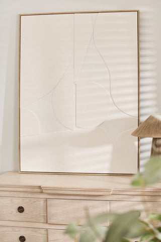 Abstract art piece on a wall above a wooden dresser with a lamp and plant.