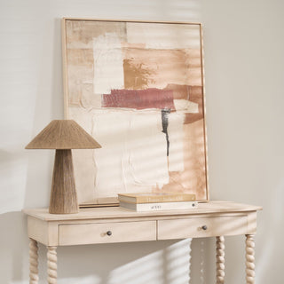 Light wooden console table with a lamp and books against a wall with abstract art.