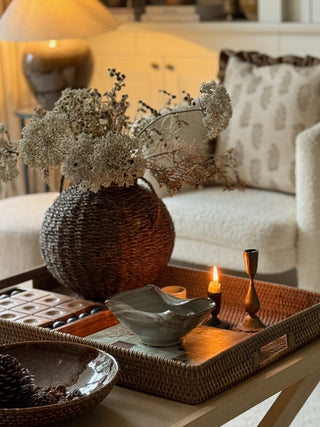 A styled interior scene with a woven brown basket vase holding dried white flowers on a tray, alongside candles, a ceramic bowl, and a sofa in the softly lit background.