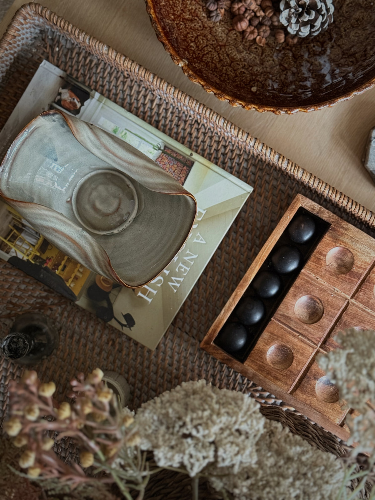 A styled flat-lay featuring a rustic ceramic pitcher with a ribbed handle and a matching ceramic bowl, arranged on a woven tray alongside a wooden game board, dried flowers, and other home decor.