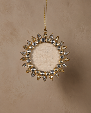A gold circular wreath ornament decorated with marquise-cut clear crystals, hanging from a gold string against a textured beige background.