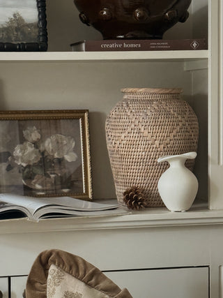 Handwoven rattan vase with rolled rim styled on a shelf beside a white ceramic jug and gilt framed print