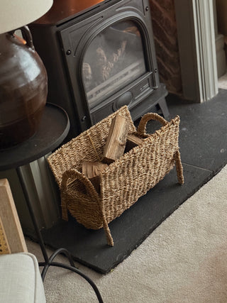 A natural woven seagrass firewood log basket with handles, filled with firewood, set on a black slate hearth in front of a black wood-burning stove.
