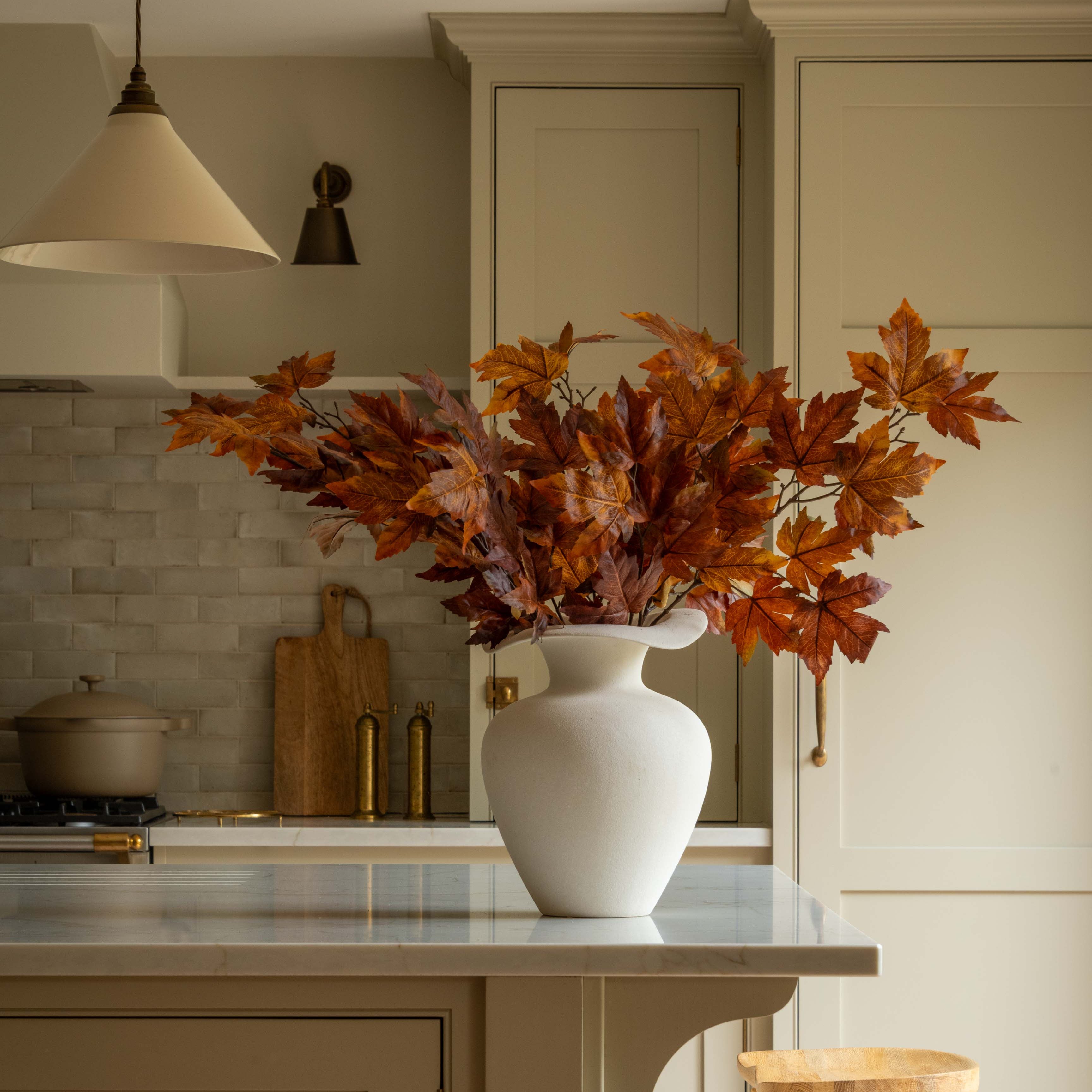 White vase with autumn leaves on a kitchen island with wooden stools.