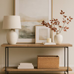 A modern console table with a light wood surface and black metal frame is styled against a neutral wall. Above it hangs a large abstract landscape artwork in soft tones with a smaller framed piece leaning against it. On the table, a ceramic lamp sits on one end, while stacked books, a candle, a candle snuffer, and a ceramic vase with autumnal stems are arranged on the other. A woven basket rests neatly on the lower shelf. The floor features pale herringbone wood with a neutral rug partially visible.