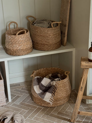 Three natural woven seagrass baskets with handles are arranged in a styled entryway. Two baskets sit on a white bench, one on the floor, with a plaid throw blanket tucked into the largest basket.