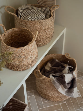 Three natural woven seagrass storage baskets with handles, displayed on a white shelf and on the floor, holding plaid blankets in a neutral-toned room.