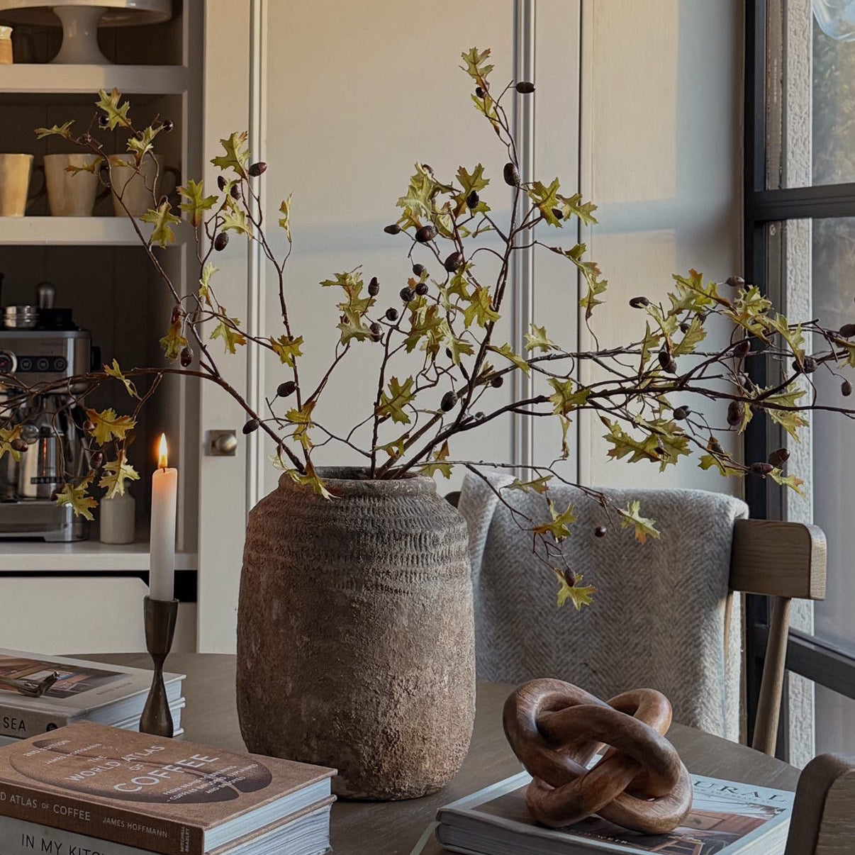 Decorative setup with branches in a vase on a table, books, and a candle in a modern interior setting.