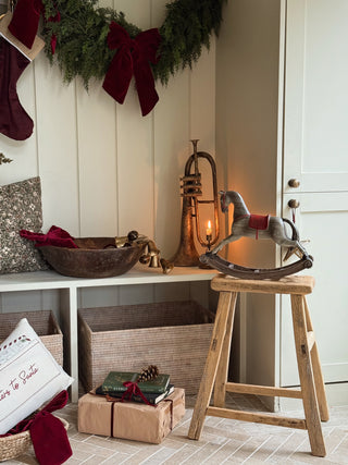A cozy holiday-themed interior scene with a rustic rocking horse on a wooden stool, a brass candle holder, wrapped gifts, woven storage bins, a green garland with a burgundy bow, and stockings hanging on a white paneled wall.