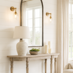 A rustic wooden console table with turned legs is placed against a white shiplap wall in a bright living room. Above the table hangs a large, black-framed arched mirror, flanked by two brass wall sconces with glass shades. On the table are a ceramic lamp with a white shade, a small bowl of greenery, and several pillar candles. Light linen curtains frame a large window, allowing natural light to fill the space.