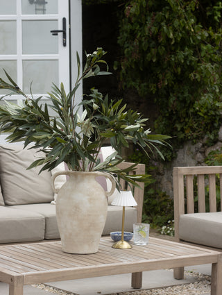 Outdoor setting with a vase of greenery on a wooden table, surrounded by beige furniture and a glass of water.