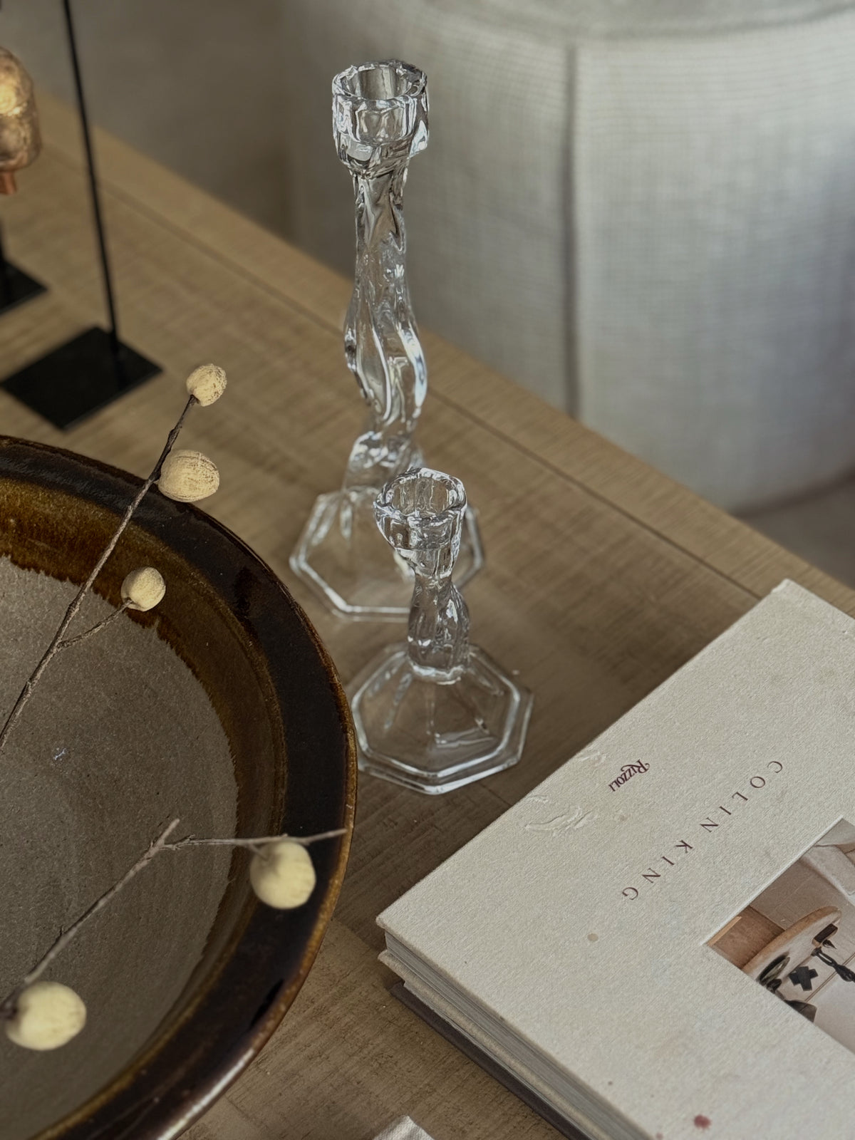 Two clear faceted glass candlestick holders on a light wood table, styled with a ceramic bowl, dried botanicals, and a book.