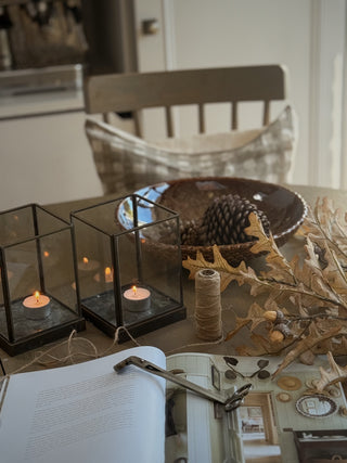 a kitchen table set with two hurricanes, oak leaves and a brown ceramic bowl