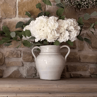 White vase with white flowers on a wooden surface against a stone wall.