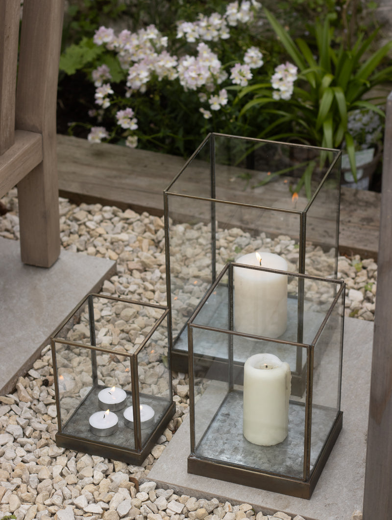 Three Whitstable bronze hurricane lanterns in small, medium and large sizes, grouped outdoors on a stone step beside a flower border, with lit pillar candles and tealights