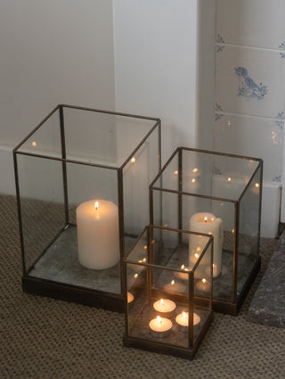Three Whitstable bronze hurricane lanterns grouped on a stone hearth indoors with foliage and lit candles, demonstrating layered styling