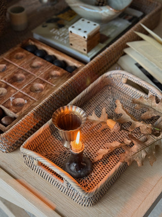 A styled autumn-themed tabletop scene featuring a woven rattan tray holding a lit candle in a dark holder and dried oak leaves, with another woven tray and wooden game pieces in the background.