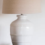 This luxuriously crafted lamp features a white ceramic base adorned with intricate beading, and accompanied by a natural linen shade.
