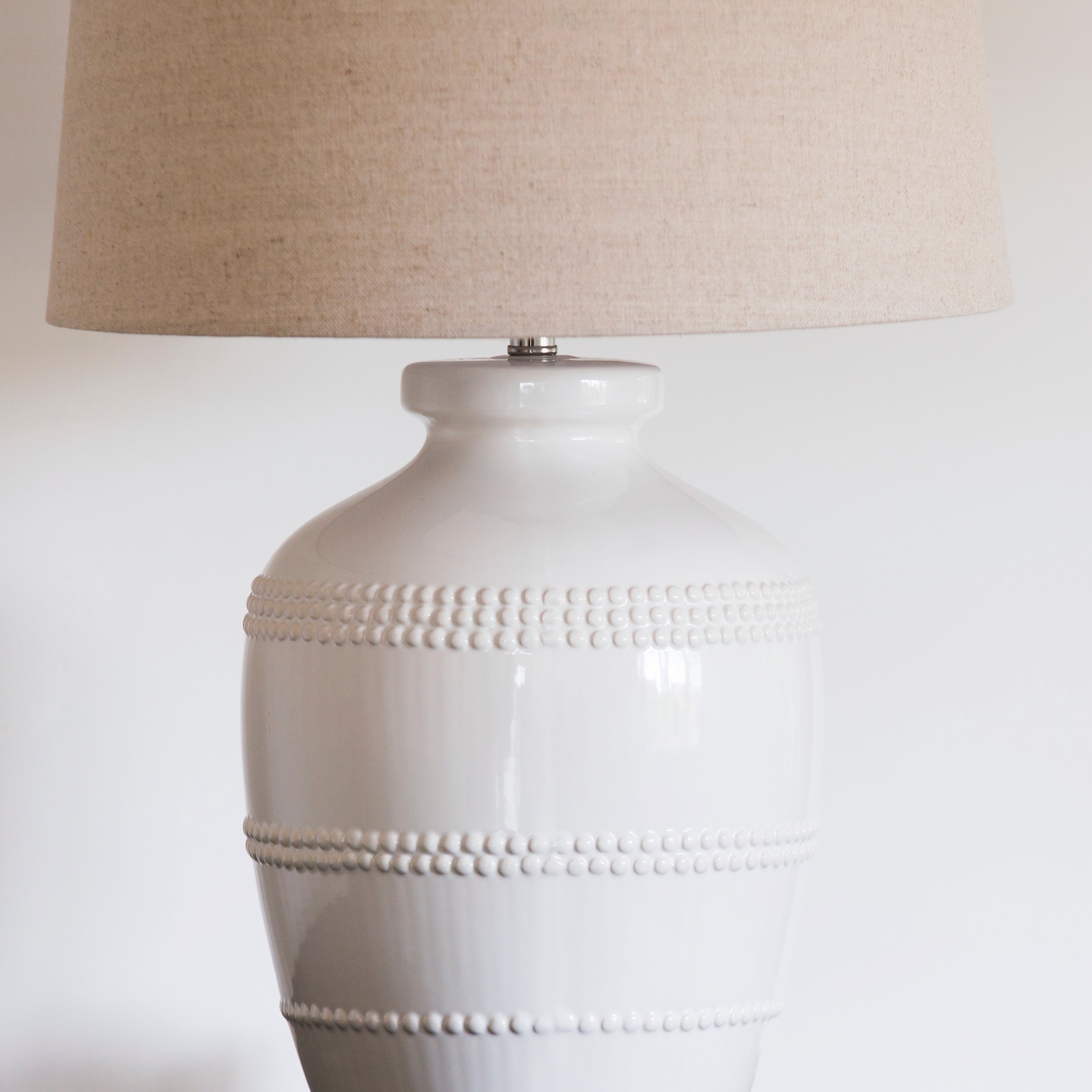 This luxuriously crafted lamp features a white ceramic base adorned with intricate beading, and accompanied by a natural linen shade.