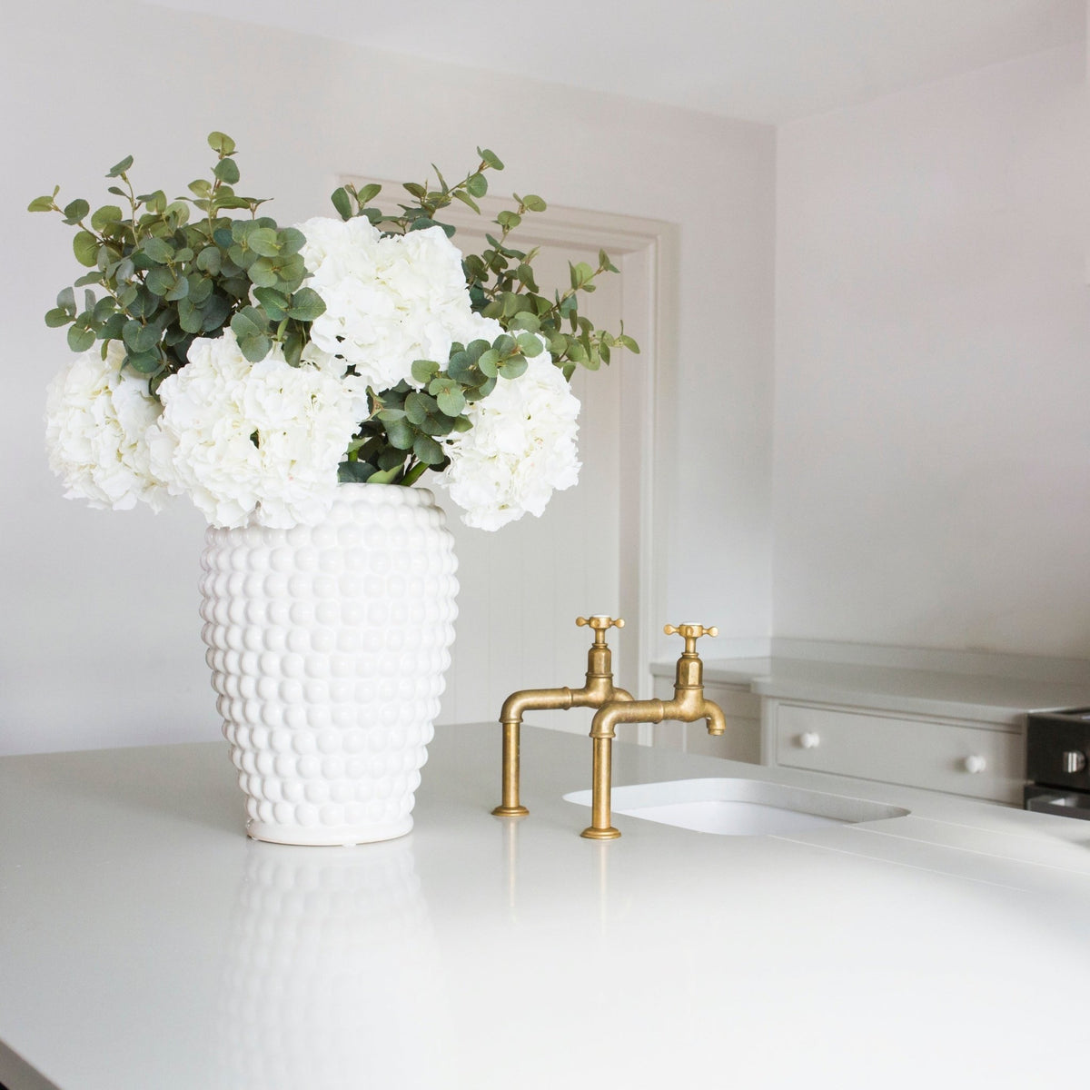 White vase with white flowers and green leaves on a kitchen counter with gold faucet.