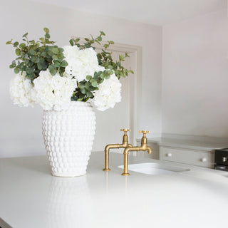 White vase with white flowers and green leaves on a kitchen counter with gold faucet.